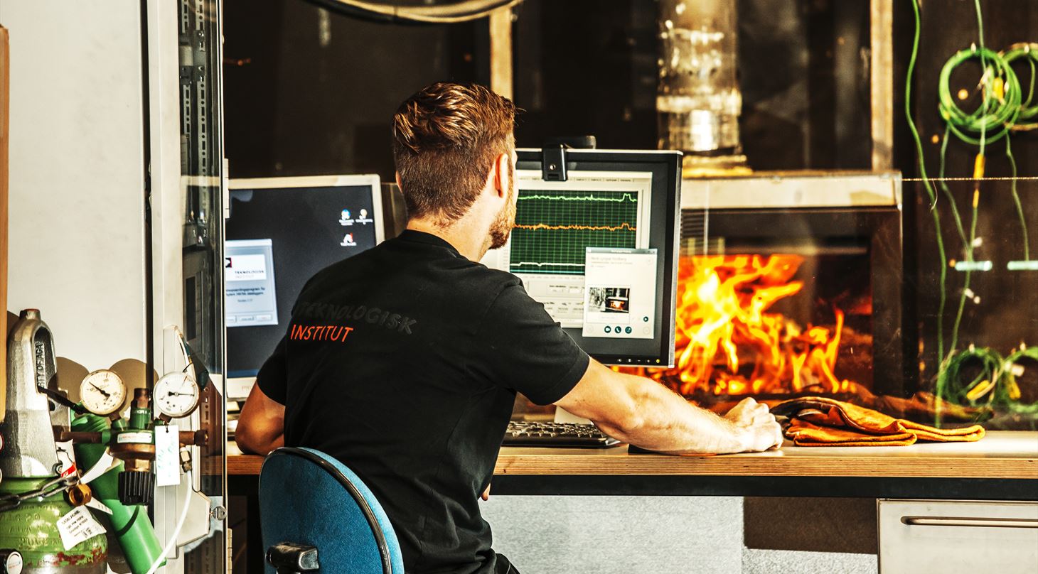 Mand i Teknologisk Institut T-shirt sidder ved et skrivebord foran en computer, hvor grafer er synlige på skærmen og arbejdshandsker liggende på bordet. I baggrunden er en tændt brændeovn samt synlige grønne ledninger.